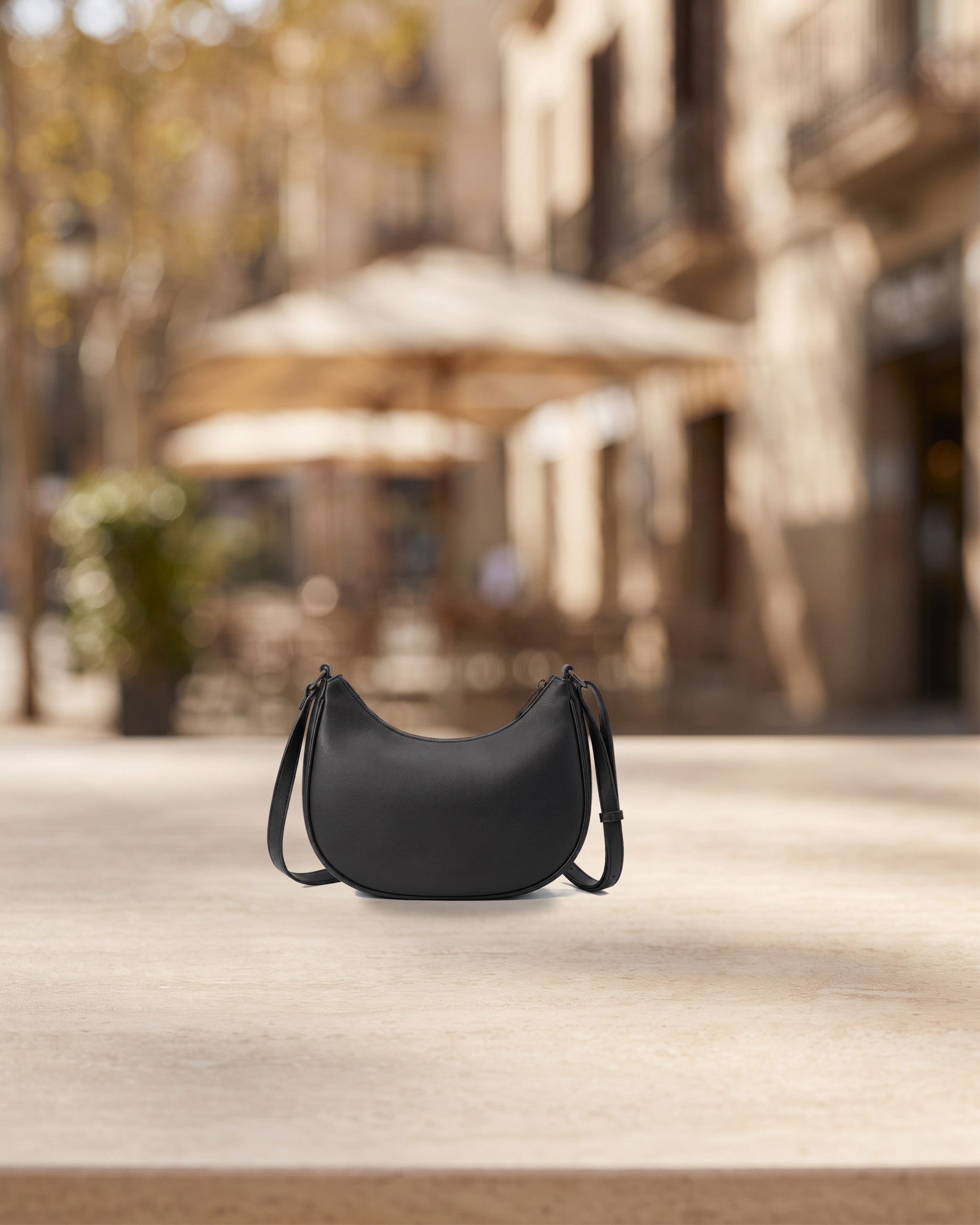 Matte black crescent-shaped shoulder bag on a pale stone surface with a blurred café street behind. Internal concept image of a fictional product.