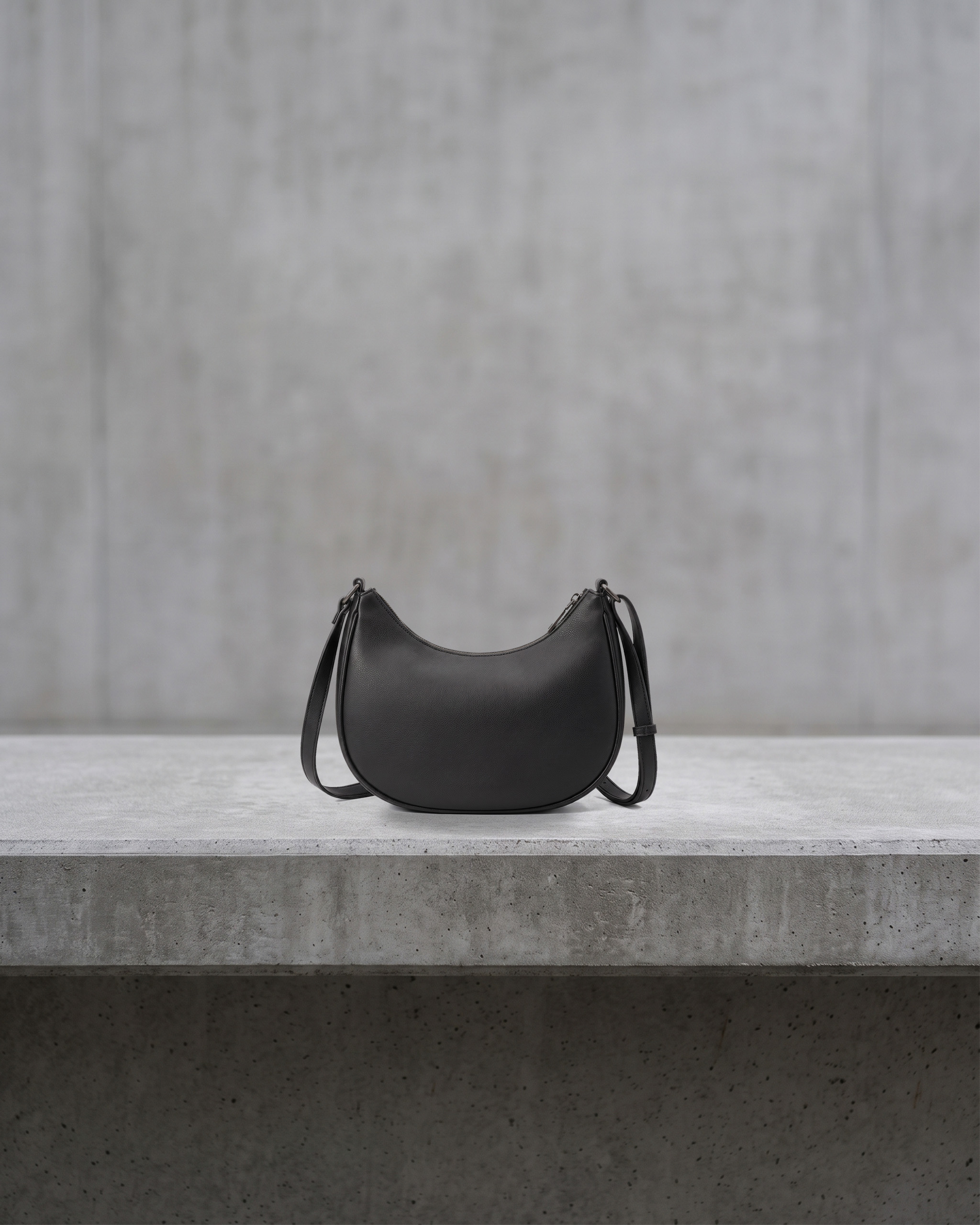 Matte black crescent-shaped shoulder bag on a flat concrete ledge in front of a grey concrete wall. Internal concept image of a fictional product.