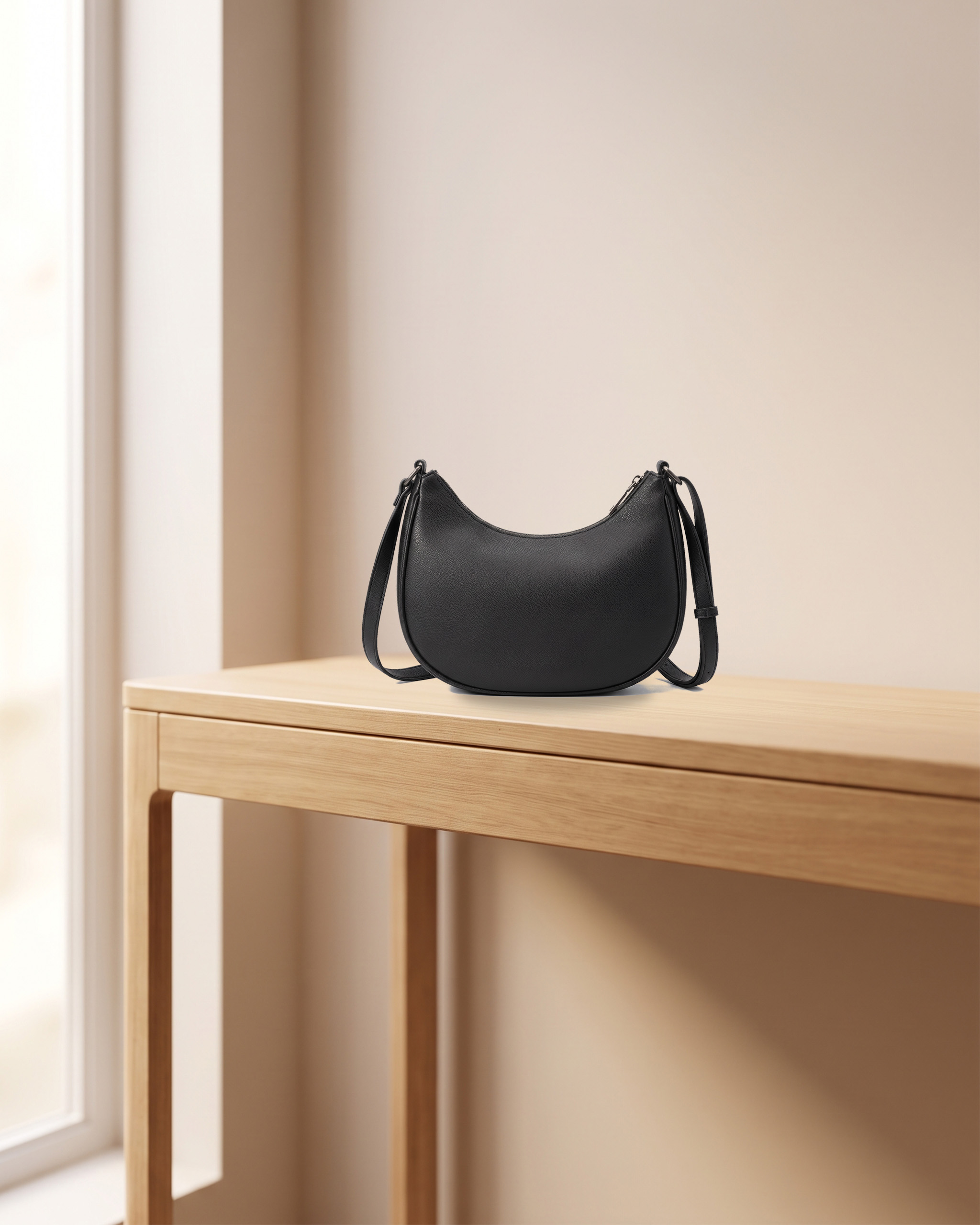 Matte black crescent-shaped shoulder bag on a light wooden console table beside a window. Internal concept image of a fictional product.